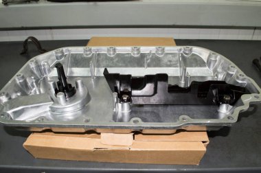 İçten yanmalı motorun yeni alüminyum yağ tavası gri bir metal çalışma masasındaki karton kutunun üzerinde duruyor.