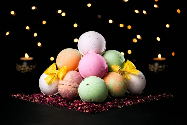 Pyramid of colorful bath bombs on the dark background surrounded by dried rose petals, candles and light in the background