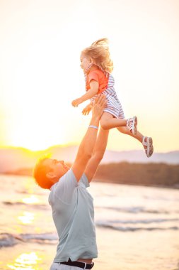 Baba ve küçük kız gün batımı kumsalında oynuyorlar, baba kızını kumsalda uçuruyor. Birlikte aile kavramını seviyoruz. Babalar Günü bayrağı