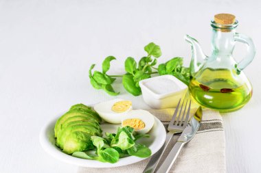 Taze avokado dilimleri, haşlanmış yumurta ve beyaz tabakta taze marul. Zeytin yağı şişesi ve masada tuz. Sağlıklı keto diyet kahvaltı. 