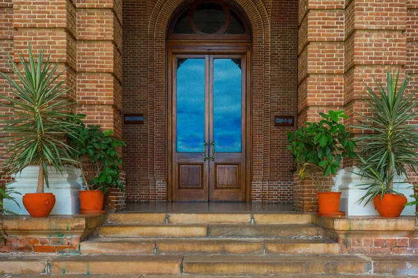 Picture of closed wooden door of a convocation hall of an institute with red bricked wall and red flower pots and stairs in the foreground. There is blue sky reflected from the glass pane on the door.