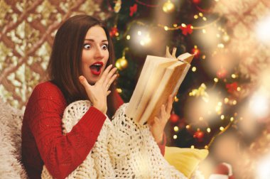 Kırmızı kazaklı güzel korkmuş kadın örgü battaniye altında koltukta oturur ve eski bir kitap okur. Çelenk içinde belgili tanımlık geçmiş Noel ağacı.