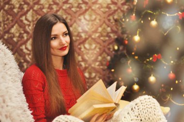 Kırmızı kazaklı genç güzel bir kadın örgü battaniye altında bir koltukta oturur ve eski bir kitap okur. Çelenk içinde belgili tanımlık geçmiş Noel ağacı.