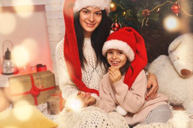 Güzel genç anne Noel Baba şapkaları kızıyla birlikte Noel ağacının altında oturan ve sarılma.
