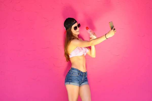 ヒップホップ スタイルの服 サングラス スマート フォンでポーズでセクシーな若い女性 水のボトルと Selfie を取る — ストック写真