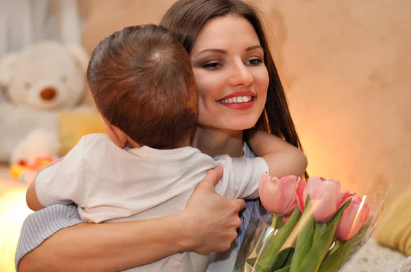 Little child son gives his mother a bouquet of delicate pink tulips and hugs her.