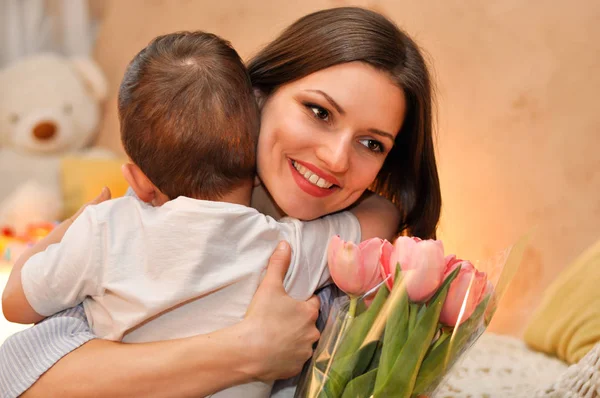 Little child son gives his mother a bouquet of delicate pink tulips and hugs her.