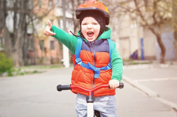 ヘルメットと赤いベストを着た小さなクールな少年は親指を上げ 背中に青いバックパックを持つバランスバイクに乗ります — ストック写真