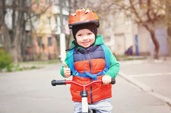 ヘルメットと赤いベストを着た小さなクールな少年は親指を上げ 背中に青いバックパックを持つランニングバイクに乗ります — ストック写真