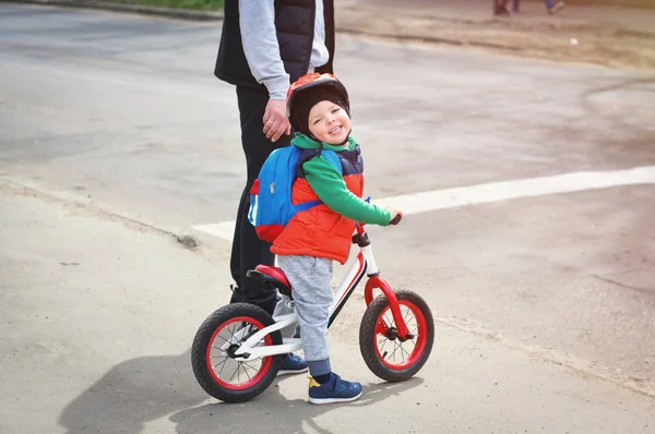 ヘルメットと赤いベストを着た小さなクールな少年は 背中に青いバックパックを背負ったバランスバイクで道路の近くに父親と一緒に立っています — ストック写真