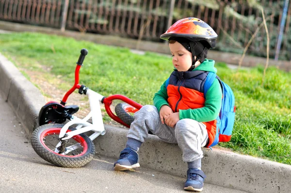 保護ヘルメットのスポーツキッドの少年は 彼の赤いホワイトバランスバイクの近くの縁石に座っています — ストック写真