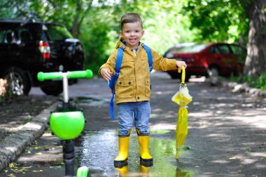 Sarı yağmurluklu küçük sevimli bir çocuk su birikintilerinde gülümseyerek ve limon rengi nde bir şemsiye tutarak yürüyor..