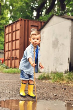 Sarı lastik çizmeler ve kot mavisi gömlek üç yaşındaki bir çocuk bir su birikintisi bir sopa ile oynar