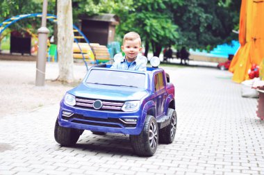 Sevimli, küçük bir çocuk bir yaz parkında elektrikli mavi bir araba sürüyor..