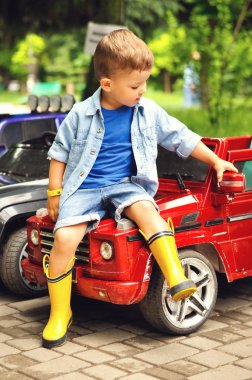Biraz sevimli üç yaşındaki çocuk sarı lastik çizmeler bir parkta kırmızı bir elektrikli arabanın kaputunda oturur.