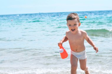 Beyaz yüzme şort plajda Sevimli küçük çocuk plastik bir sulama ile dalgalar üzerinde çalışır olabilir ve gülüyor.