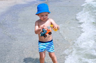 Sahilde mavi çizgili yüzme şort sevimli bir küçük çocuk bir oyuncak ekskavatör ile oynar.
