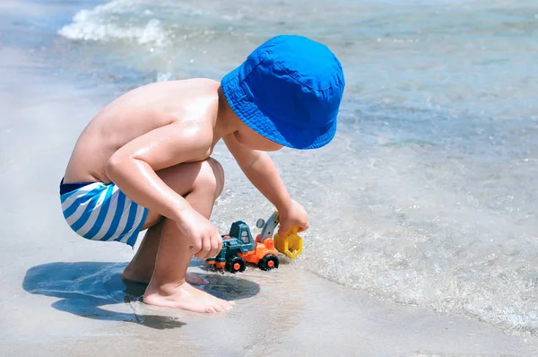 Mavi çizgili mayolu şirin bir çocuk kumsalda oturup oyuncak kazıcısıyla oynuyor..