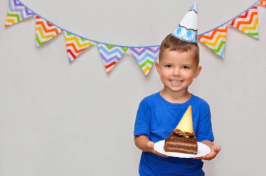 Küçük bir çocuk şenlikli bir şapka içinde gülümsüyor ve çikolatalı kek bir parça tutar. Doğum gününü kutluyor.