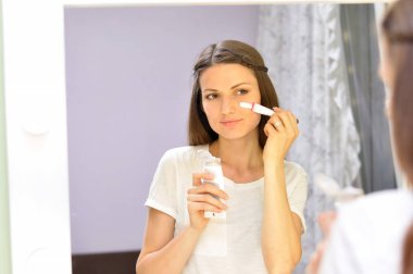 Genç bir güzel kadın beyaz bir t shirt soyunma odası aynaönünde evde dururken onu temizlenmiş yüzüne beyaz bir maske koyar.