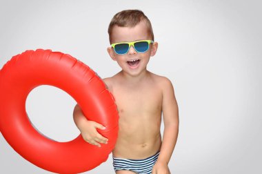 Sevimli bir küçük çocuk kırmızı şişme halka ve yeşil güneş gözlüğü ile çizgili yüzme şort gülüyor.