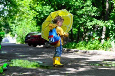 Sarı yağmurluklu küçük sevimli bir çocuk su birikintilerinde gülümseyerek ve limon rengi nde bir şemsiye tutarak yürüyor..