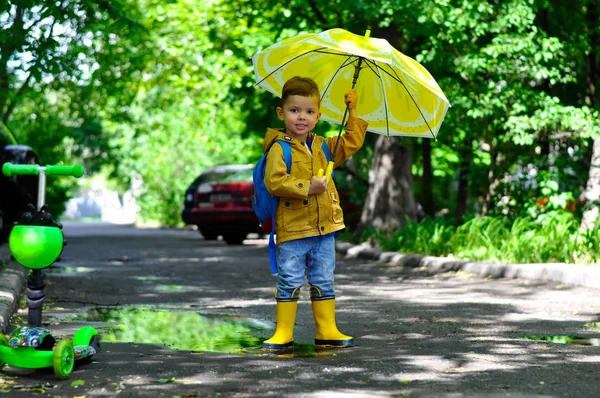 黄色のレインコートを着た小さなかわいい少年が レモンの色で傘を持って微笑みながら水たまりを歩く — ストック写真