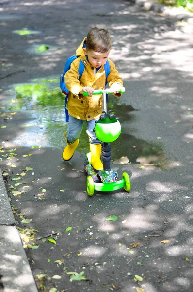 黄色いレインコートを着た小さなかわいい少年で 背中にバックパックが付いているかわいい少年は 緑のスクーターの水たまりを乗り切る — ストック写真