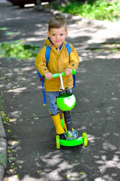 黄色いレインコートを着た小さなかわいい少年で 背中にバックパックが付いているかわいい少年は 緑のスクーターの水たまりを乗り切る — ストック写真
