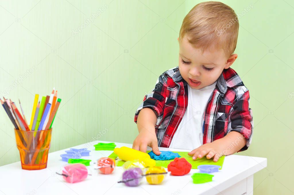 Happy boy playing with plasticine. A two-year-old baby boy in a red checked shirt sits at a table