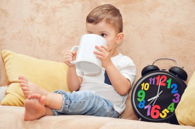 Beyaz tişörtlü şirin bir çocuk evdeki bir kanepede oturur ve büyük beyaz bir bardaktan çok renkli bir çalar saatin yanında su içer..