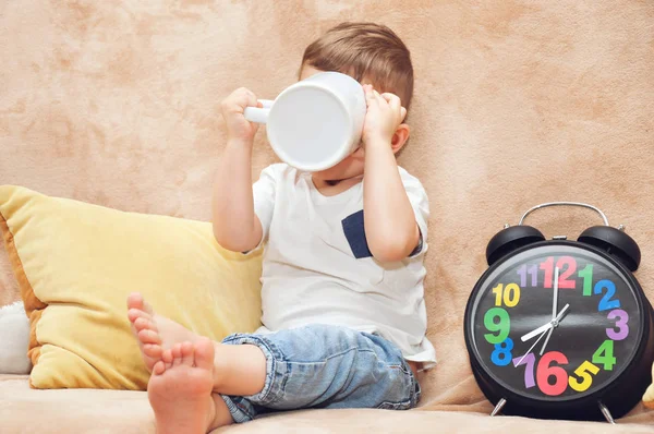 Beyaz tişörtlü şirin bir çocuk evdeki bir kanepede oturur ve büyük beyaz bir bardaktan çok renkli bir çalar saatin yanında su içer..