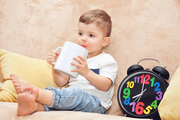 Beyaz tişörtlü şirin bir çocuk evdeki bir kanepede oturur ve büyük beyaz bir bardaktan çok renkli bir çalar saatin yanında su içer..