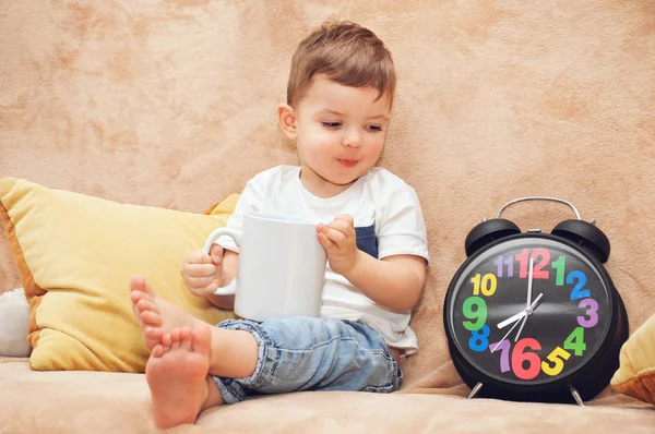 Beyaz tişörtlü sevimli çocuk elinde büyük beyaz bir fincan ile kanepede oturuyor.