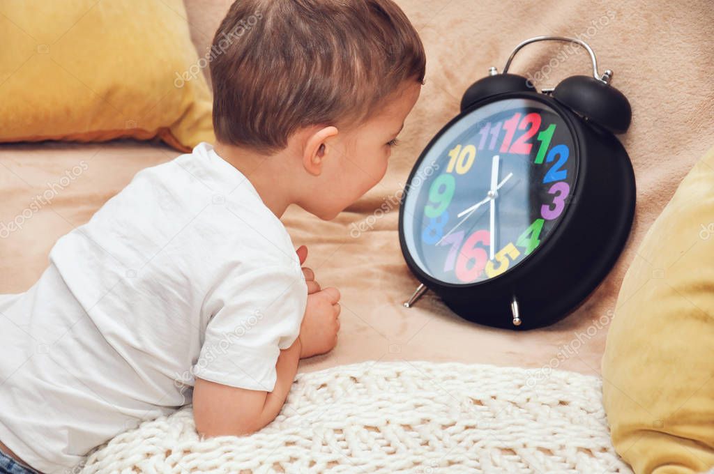 Cute little boy looks at a big black alarm clock with multi-colored numbers.