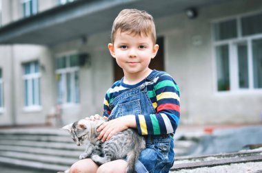 Kot pantolon giymiş şirin bir çocuk. Tulum giymiş. Diz çöküp gülümseyen bir kedi yavrusu tutuyor.