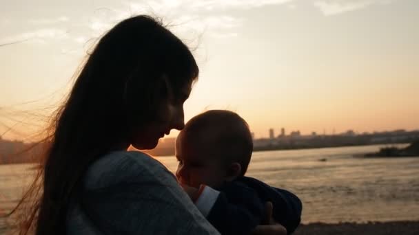 Молодая мать осторожно целует своего маленького сына, стоя у реки на закате . — стоковое видео