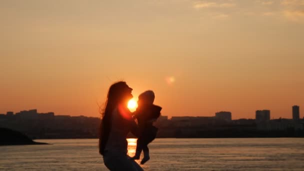 一位年轻的母亲高兴地把她的小儿子抱起来。母亲和孩子在日落时在河边散步很开心. — 图库视频影像