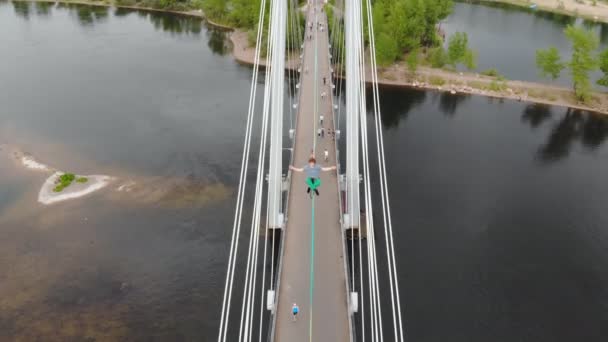 De man zit in een Lotus positie op een touw gespannen tussen de steunen van de brug op grote hoogte. Slackliner doet yoga. — Stockvideo