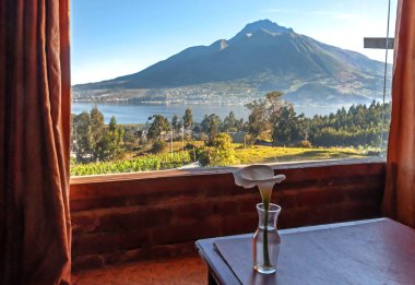 Güzel bir sabah Imbabura yanardağ ve San Pablo gölü görünümü, bir kabin odasının içinden, bir masa üzerinde ön planda bir çiçek ile. Ekvador