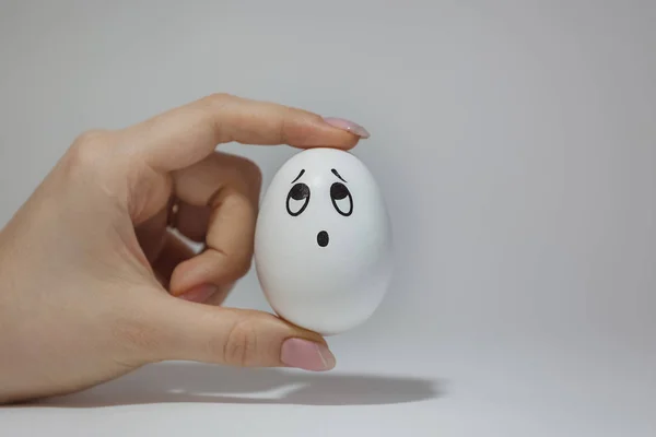 egg with a sad painted face between fingers, looks up, emotions on the egg, white background