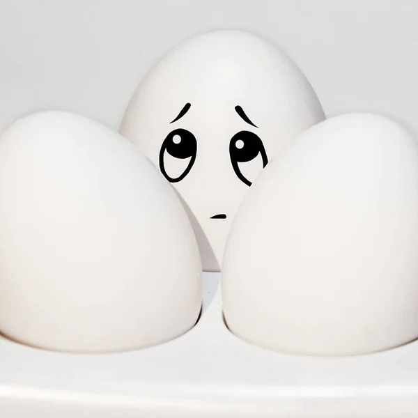 egg with a sad painted face between two white eggs, looks up, emotions on the egg, white background