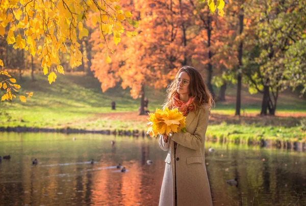 一位年轻女子走在秋天的公园里 她站在湖边 黑发女人穿着一件灰色的外套 她手里拿着一束黄叶 — 图库照片
