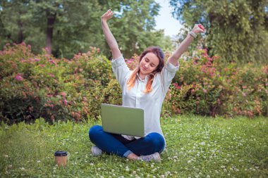 bir dizüstü bilgisayar ve kahve bir fincan ile bir kız çim, gülümsüyor ve gülümsüyor, iyi haber, çalışmanın sonu parkta oturur