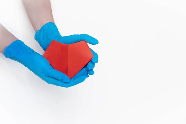 hands in blue gloves holding a red paper heart on a white background, concept of health, medicine, heart disease