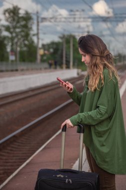 kız platformda duruyor, treni bekliyor, telefona bakıyor, programa, evden ayrılışa, bir bavula.