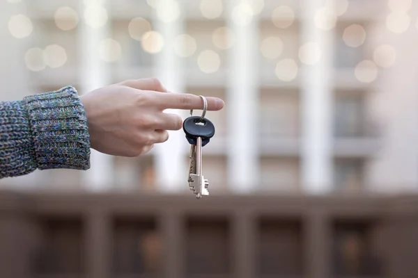 the keys to the apartment on the finger, the hand holds the keys on the background of a new high-rise building, the concept of buying or selling real estate, real estate agency