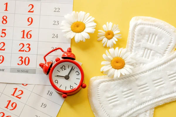 calendar, alarm clock and women's pads on a yellow background, concept of women's critical days, hygiene and women's health care
