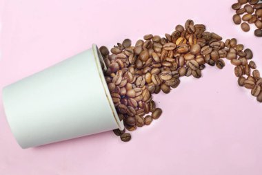 Kavrulmuş koyu kahve çekirdekleri güzel beyaz kağıt bardak bir masada izole pembe bir arka plan ile dökülür