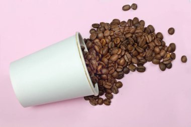 Kavrulmuş koyu kahve çekirdekleri güzel beyaz kağıt bardak bir masada izole pembe bir arka plan ile dökülür